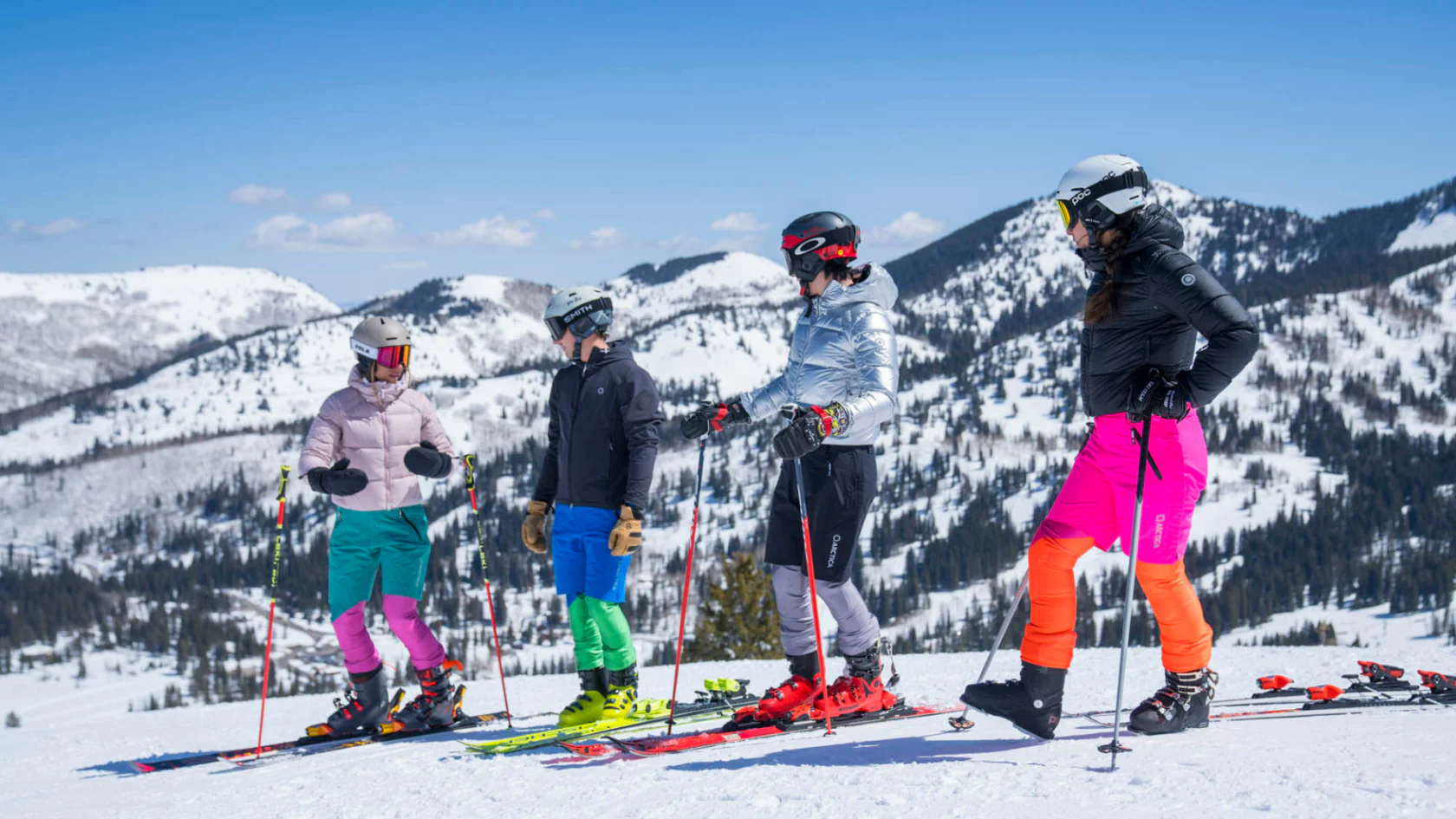 Four skiers in multicolor Arctica ski race suits, down jackets and training shorts on a snowy mountain ridge