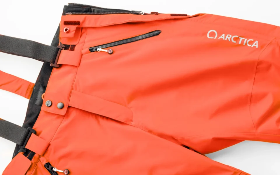 Close-up of the Arctica Side Zip Ski Pants 2.0 in Tangerine showing the waistband, suspenders, zip pocket and logo.
