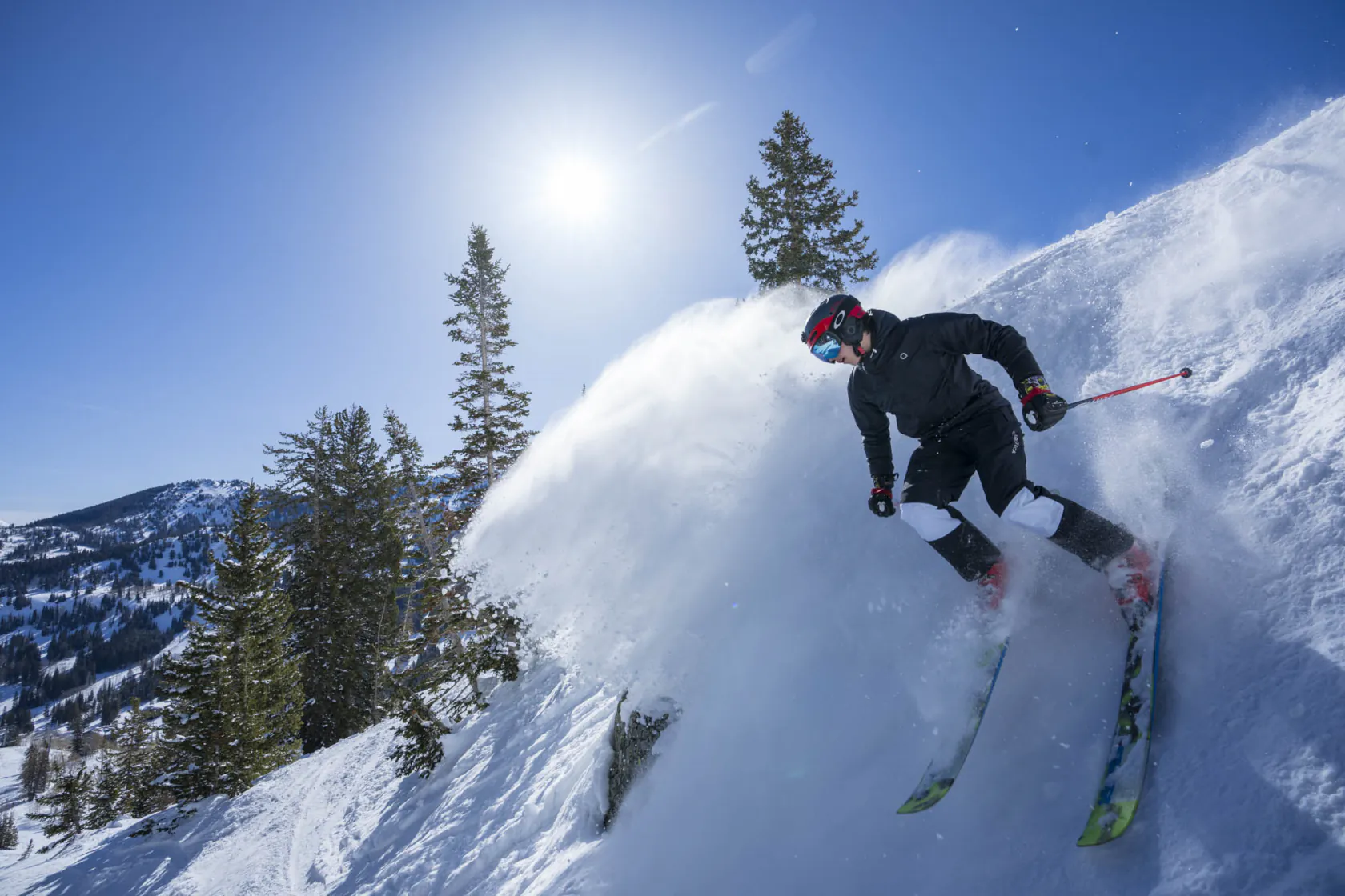 Skier in a black Arctica jacket and ski pants spraying powder down a sunny mountain slope