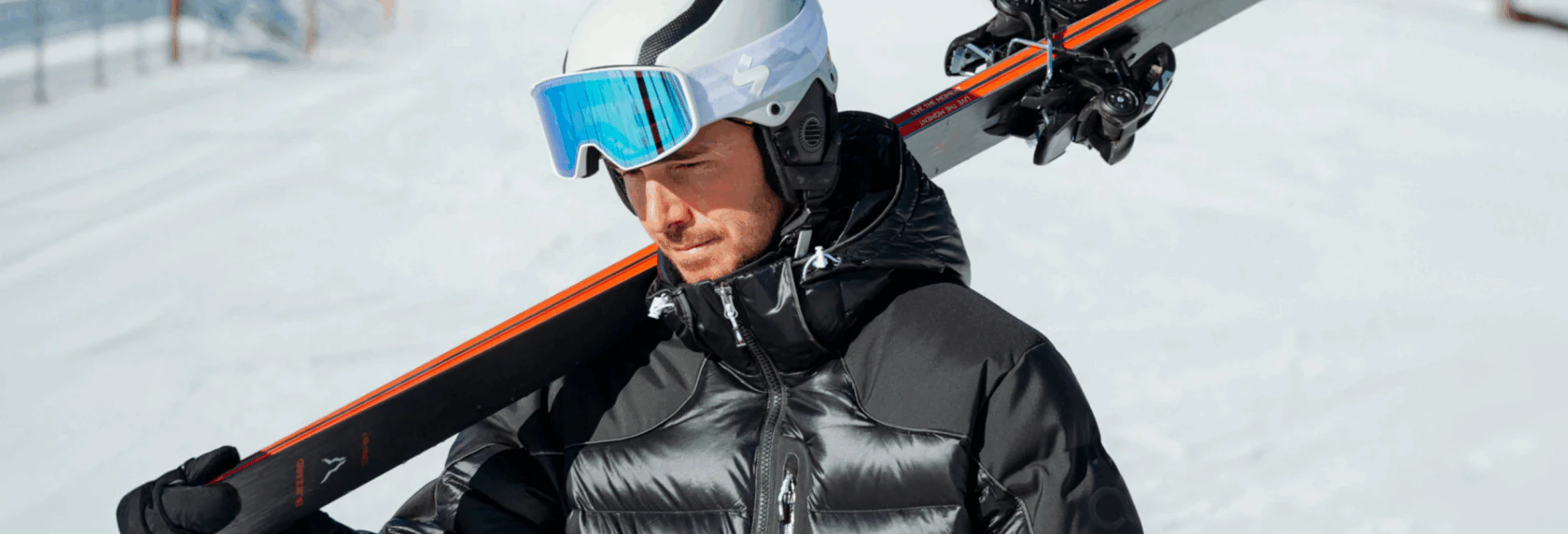 Man in a black Arctica down jacket and white helmet carrying skis on his shoulder