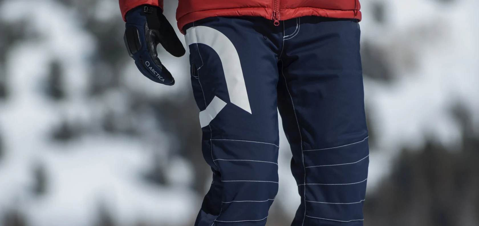 Close-up of navy Arctica side zip ski pants with a large white logo, worn with a red puffer jacket and Arctica gloves