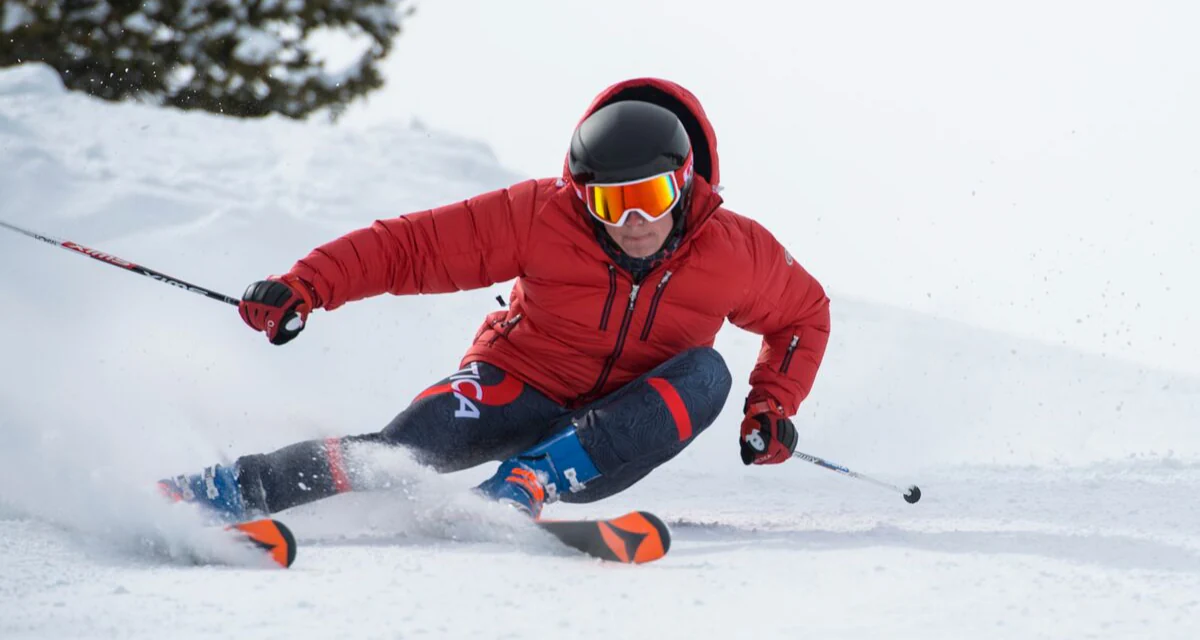 Skier in a red hooded down jacket and navy Arctica ski race suit carving on a groomed slope