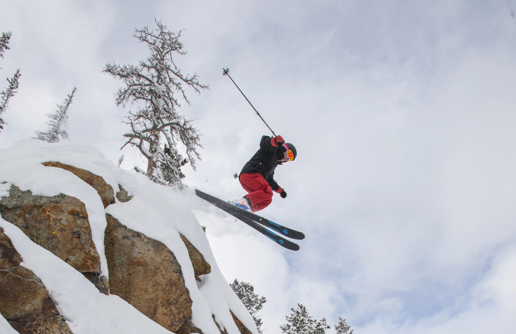 Skier in a black jacket and red Arctica side zip ski pants jumping off a snowy rock cliff