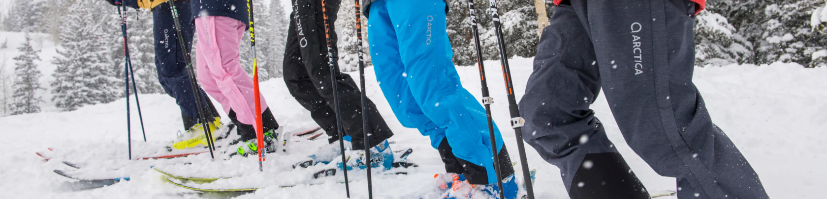 Row of skiers in navy, pink, black, blue and grey Arctica side zip ski pants in falling snow