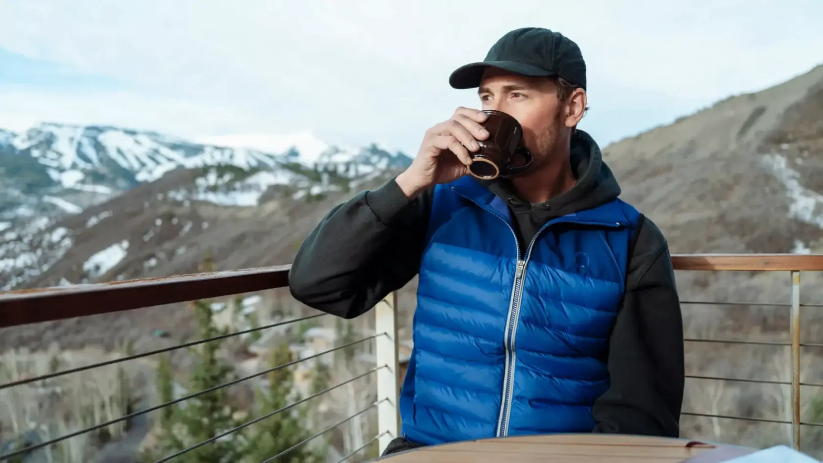Man wearing a blue Alpine Envoy Down Vest on a mountain balcony while drinking coffee.